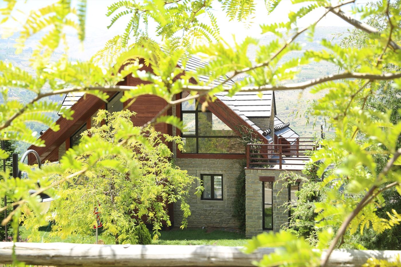 A rust-patinated steel fire bowl on black legs standing on clover-flecked lawn, with a pale stone facade and natural-wood front door reached by a low stone step beyond, framed by a large fir and overhanging green foliage in dappled sunlight.