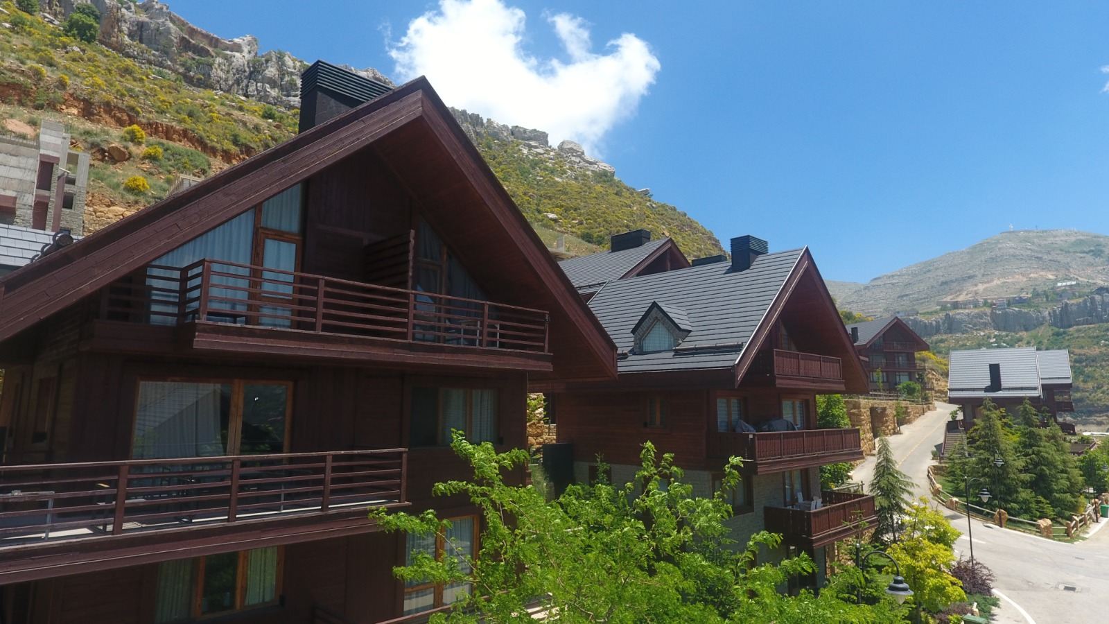 A row of dark-timber chalets with slate-grey pitched roofs and slatted balconies stepping up a curving road, with a bare rocky crag and scrub-covered mountain slope rising behind them under a blue sky.