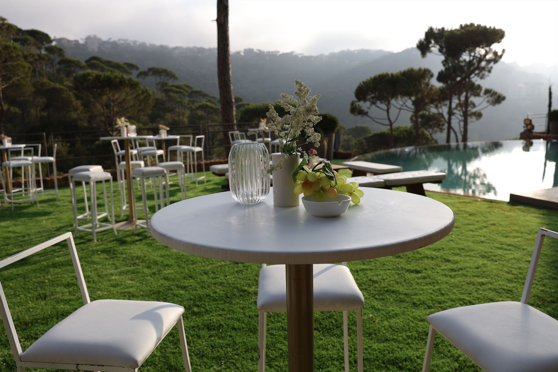 A round table holding a fluted glass vase, a white jug of dried flowers and a bowl of fruit, with white chairs on clipped lawn beside the dark water of an infinity-edge pool, and layered ridges of umbrella pines fading into haze under a low backlit sky.